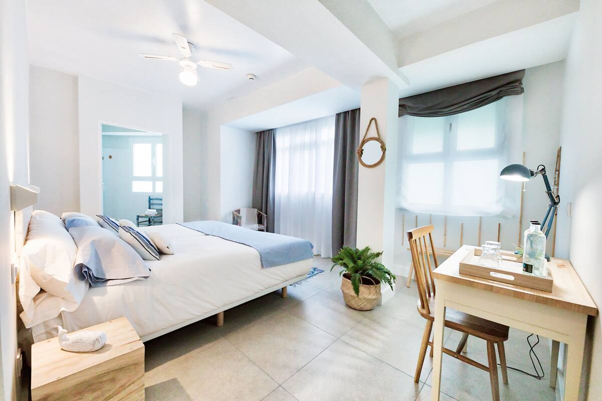 A modern bedroom with a bed, bedside table, desk, chair, and a potted plant.
