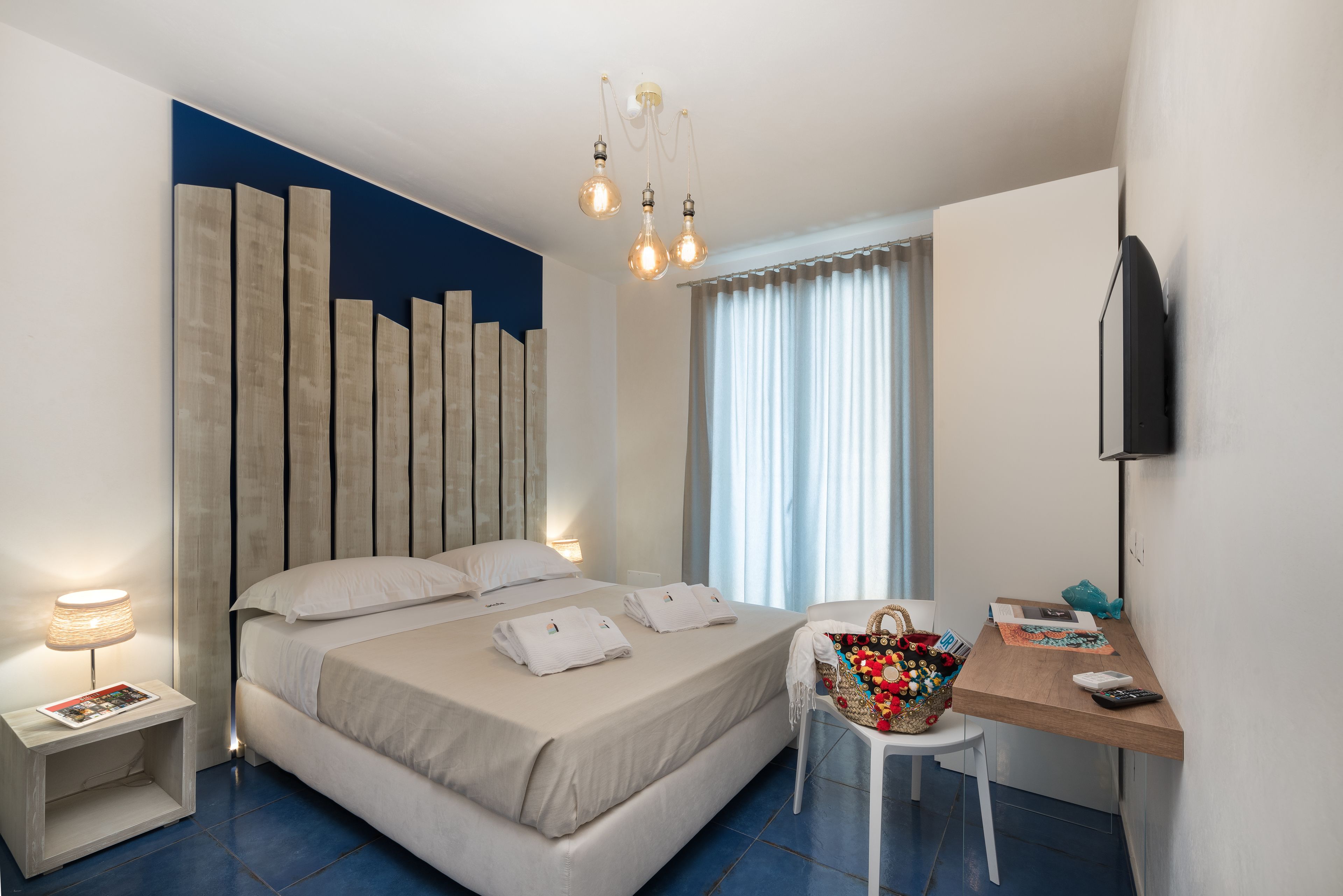 A modern hotel room with a bed, bedside table, desk, and a large window with curtains.