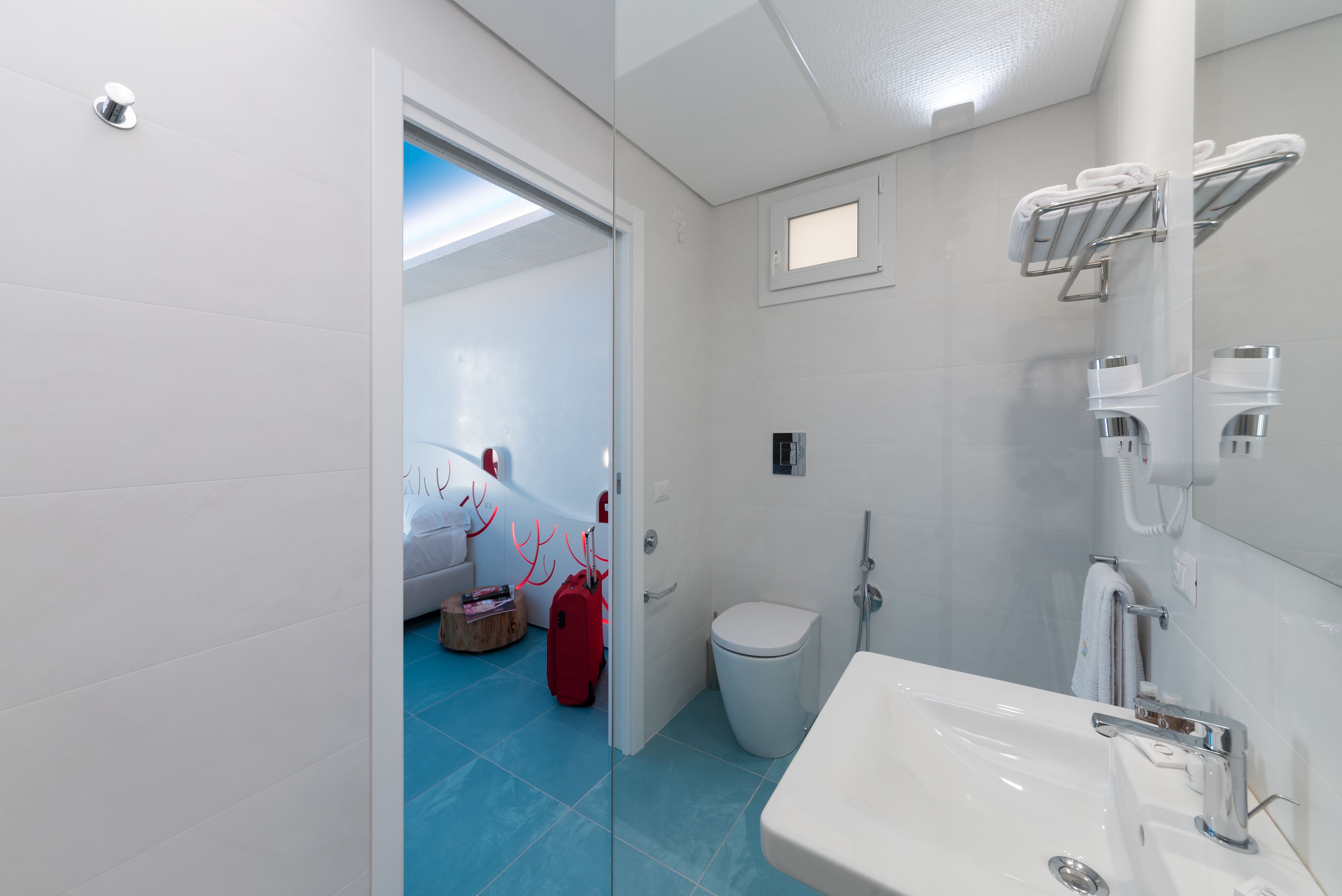 A modern bathroom with a white sink, a toilet, and a shower area. There is a glass door leading to a bedroom with a bed and a red suitcase.