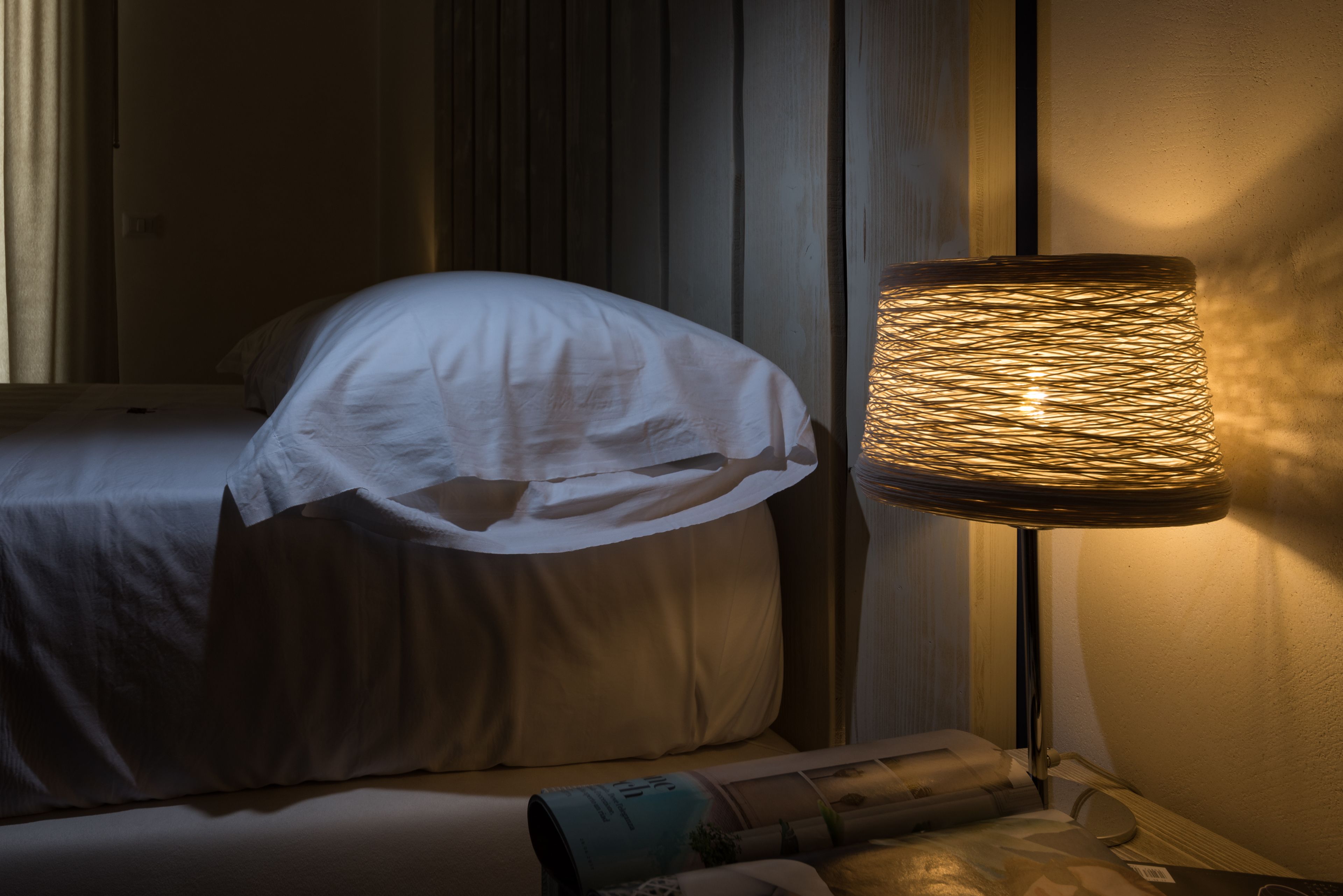 Une chambre d’hôtel faiblement éclairée, avec un lit recouvert de draps blancs, une lampe dotée d’un abat-jour en osier et un magazine posé sur la table de chevet.
