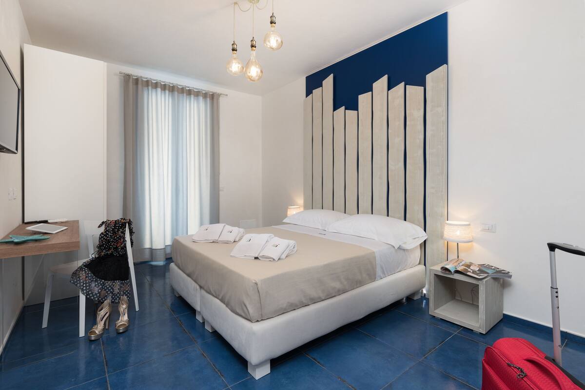 A hotel room with a bed, a desk, a chair, a suitcase, and a lamp.