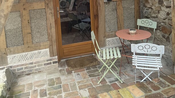 Una habitación rústica de madera con techo inclinado, una pared de piedra y una puerta de madera que da a un espacio de almacenamiento. Hay una pequeña mesa redonda con una silla y un banco con respaldo.