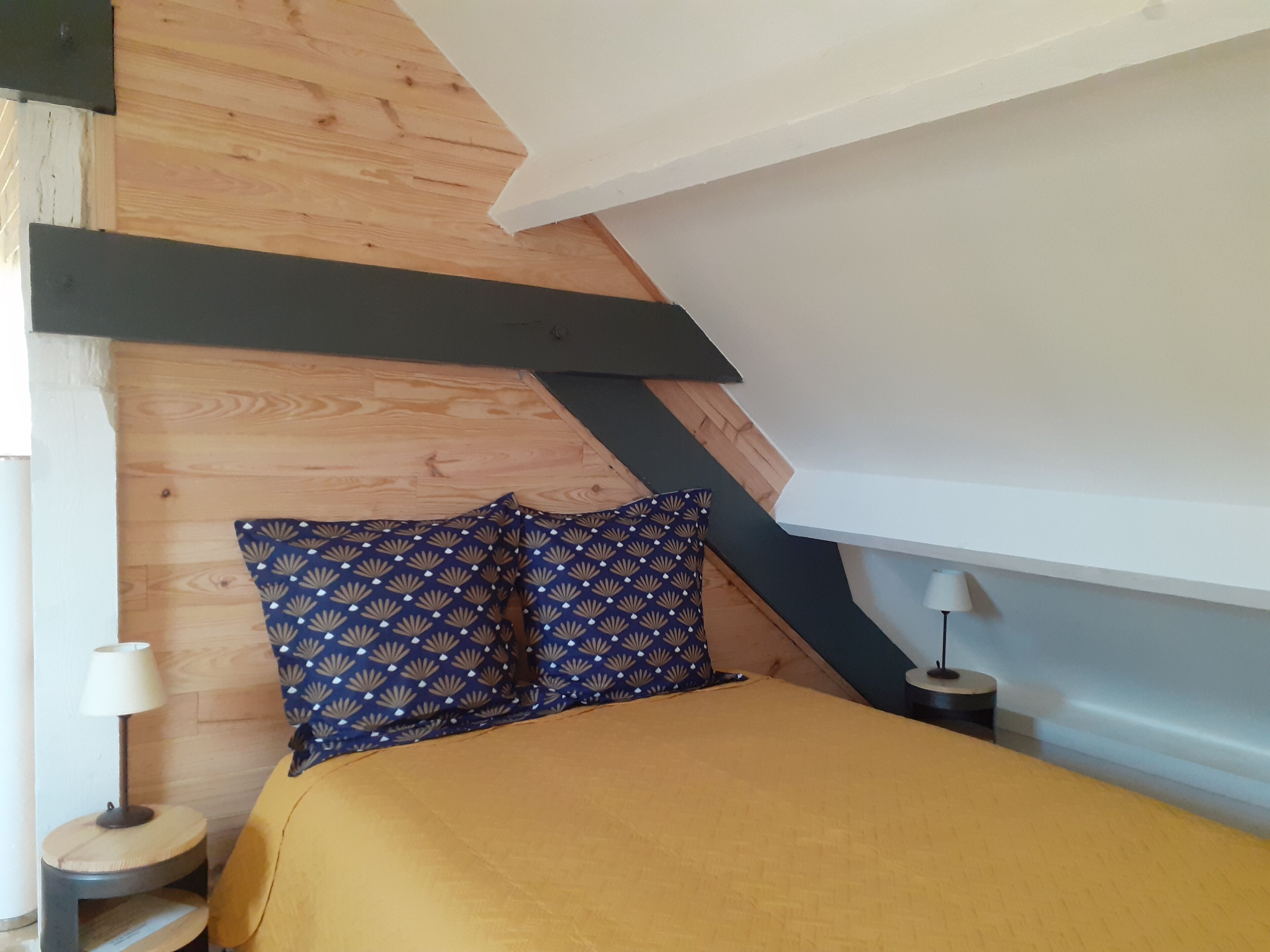 A bedroom with a sloped ceiling, wooden paneling, a bed with a yellow bedspread, two patterned pillows, a bedside lamp, and a small round table with a lamp.
