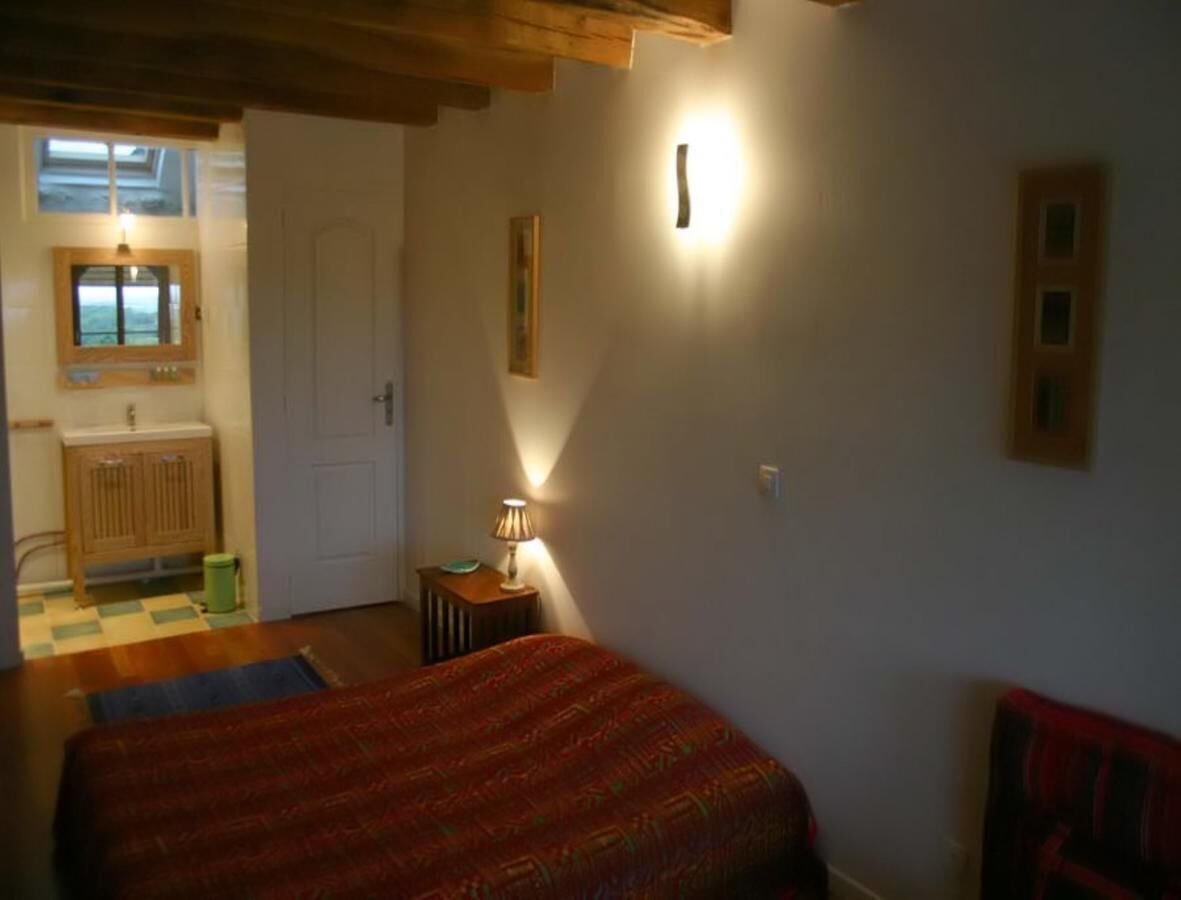 A bedroom with two beds, a wooden ceiling, a small table with a lamp, and a view of a bathroom through an open door.