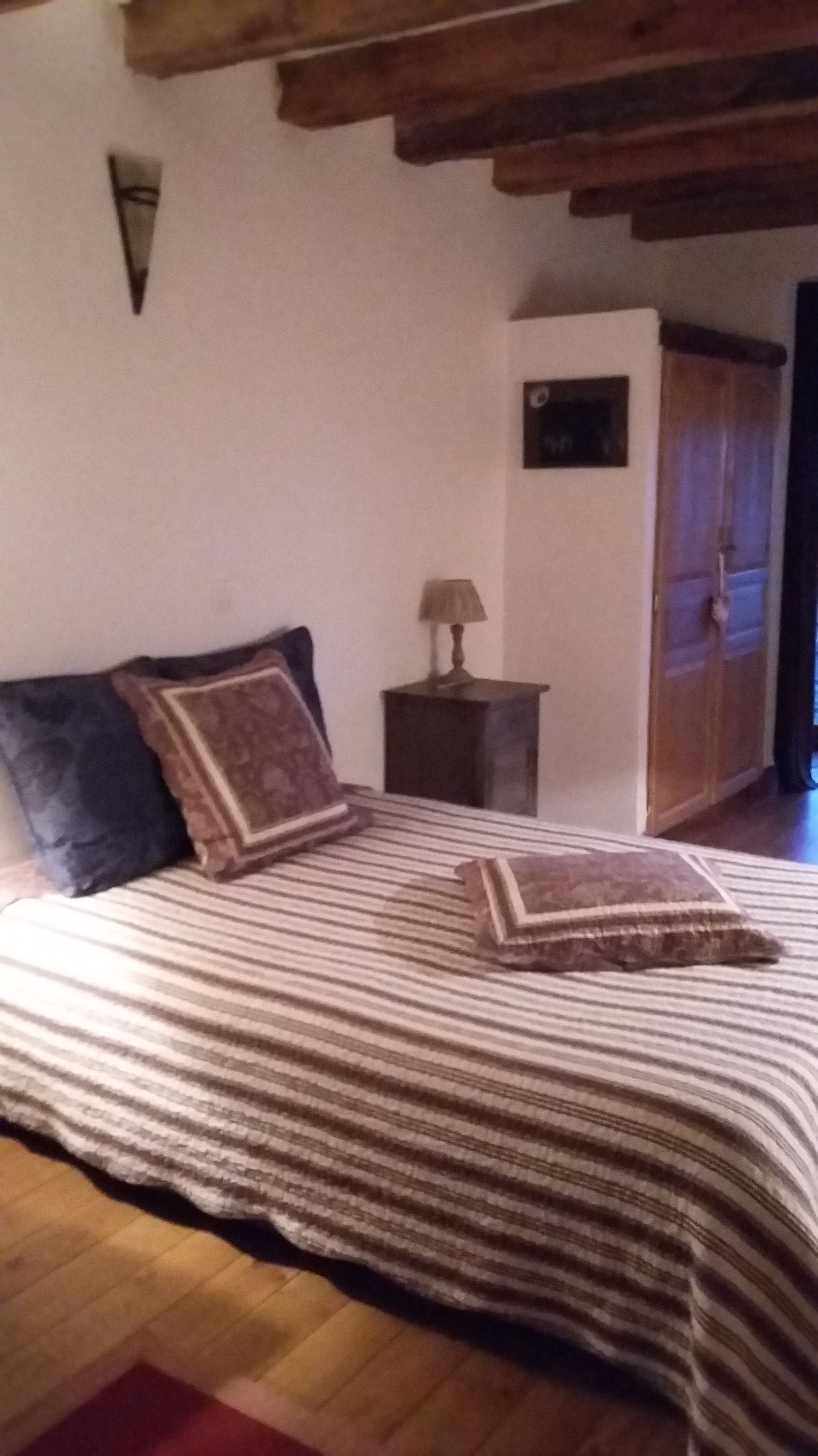 A bedroom with a wooden ceiling, a bed with striped bedding, two pillows, a nightstand with a lamp, and a wardrobe.