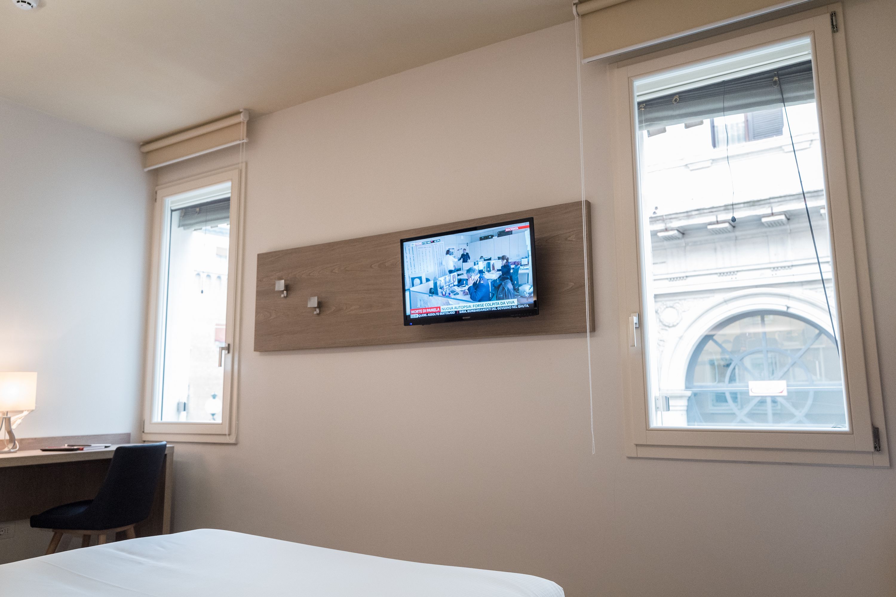 Habitación de hotel con cama, escritorio con silla, televisor montado en la pared y ventana con vista a edificios.