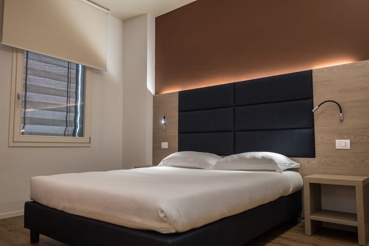 Une chambre moderne avec un grand lit, une tête de lit dotée d’un éclairage intégré, une table de chevet et une fenêtre avec des stores.