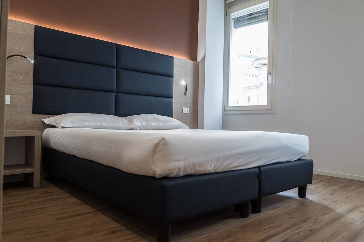 Une chambre moderne avec une tête de lit sombre, une table de chevet et une fenêtre donnant sur des immeubles.