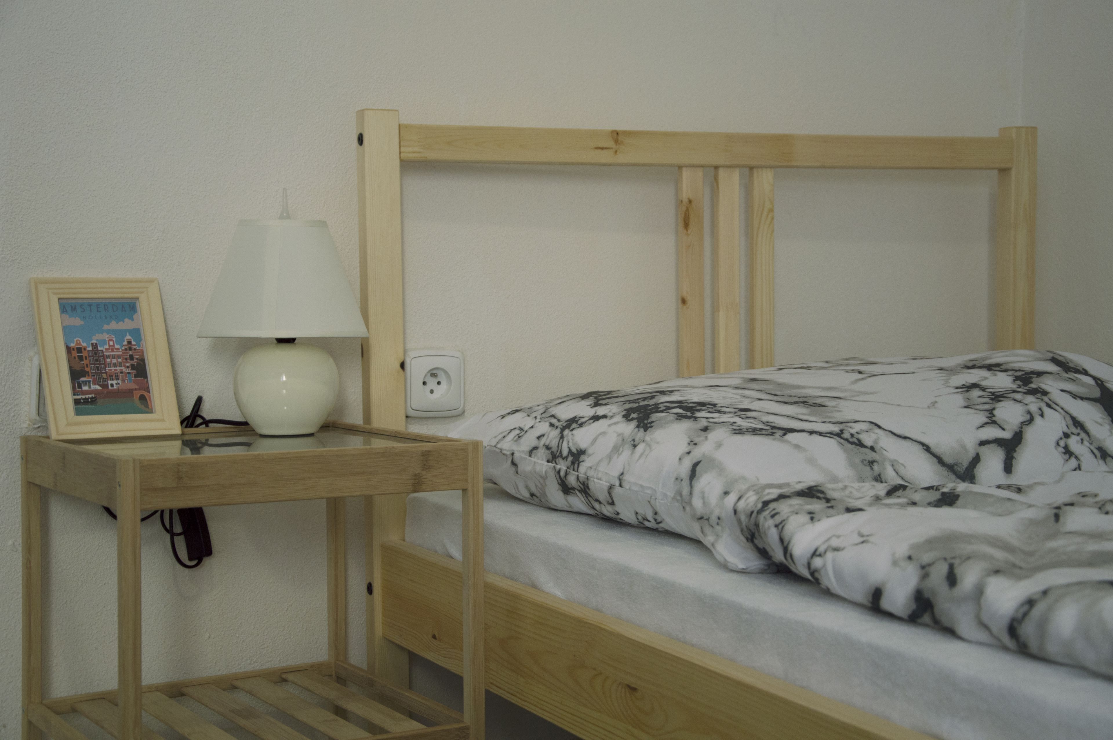 Una cama litera de madera con colchón blanco y ropa de cama con estampado blanco y negro. Hay una pequeña mesita de noche de madera con una lámpara blanca y un cuadro enmarcado.