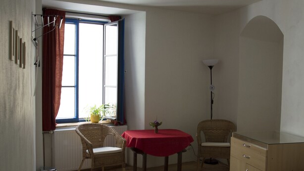 A bedroom with a bed, a small table with a red tablecloth, a wicker chair, a radiator, and a window with curtains.