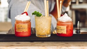 Un bar con tres bebidas sobre una bandeja, una persona sirviendo una bebida y un fondo desenfocado.