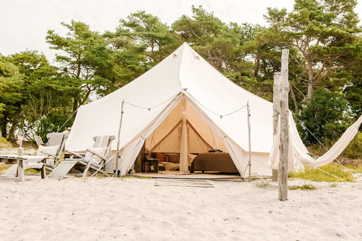 A large white tent with a pitched roof, surrounded by beach chairs and a hammock, set on sandy ground with trees in the background.