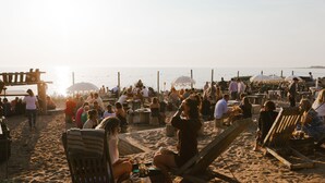 Una playa concurrida, con gente sentada en sillas y sombrillas, estructuras de madera y una extensa orilla de arena.
