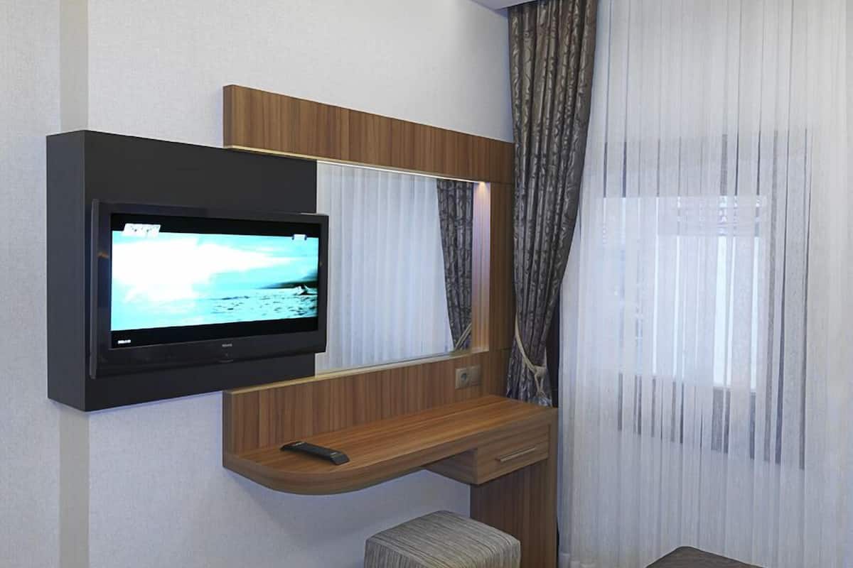A hotel room with a flat-screen TV mounted on the wall, a wooden desk, a small ottoman, and a bed with a patterned bedspread.