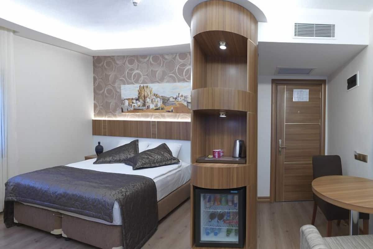 A modern hotel room with a bed, a built-in mini-fridge, a dining table, and a door with a sign.
