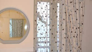 A room with a round mirror on the wall, a window with patterned curtains, and a view of the outside.