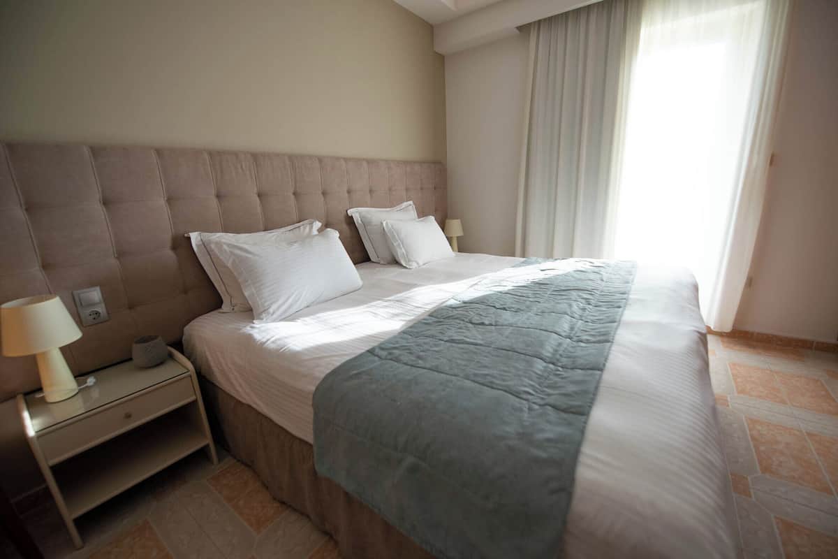 A neatly made bed with a grey blanket, a bedside table with a lamp, and a window with curtains.