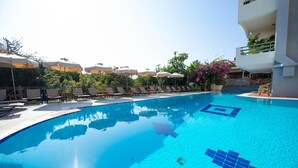 A swimming pool with lounge chairs and umbrellas, surrounded by greenery and a building with balconies.