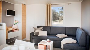 Ein modernes Wohnzimmer mit grauem Sofa, einem flachbildfernseher an der Wand, einem kleinen Beistelltisch mit Buch und Blumenvase sowie einem Fenster mit Vorhängen.