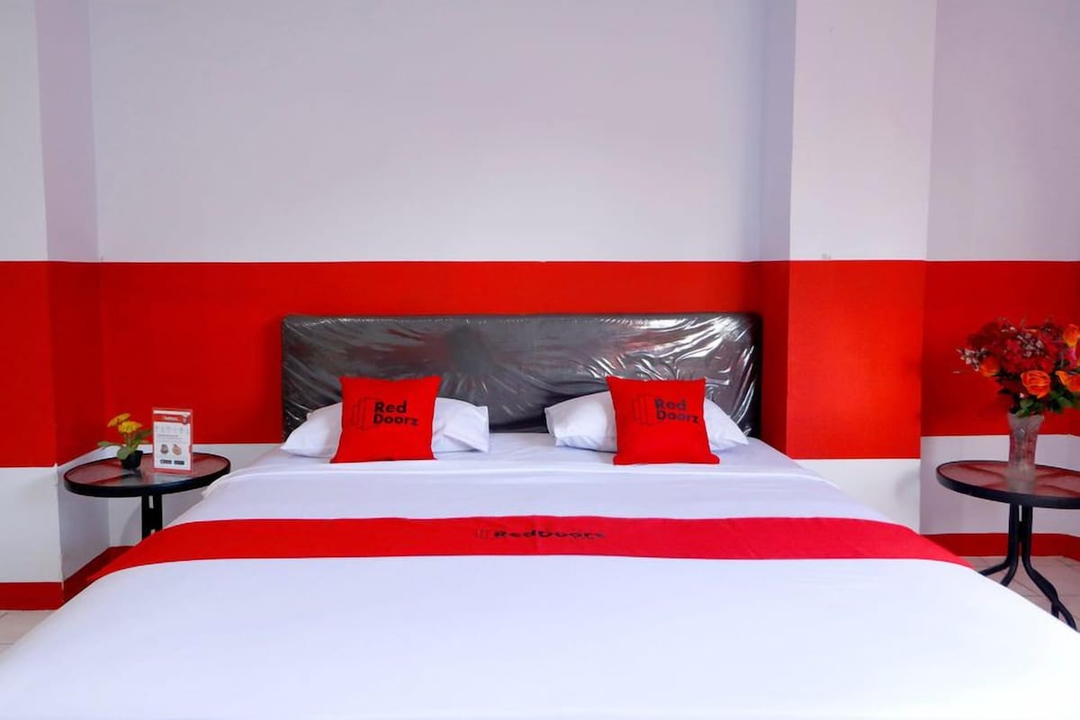Une chambre d’hôtel avec un lit, deux oreillers, une table de chevet, une petite table et un vase avec des fleurs.