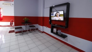 A room with a tiled floor, a small table with two chairs, a potted plant, a wall-mounted TV, and a shelf with bottled water. - RedDoorz Plus @ Samratulangi Manado (Manado)