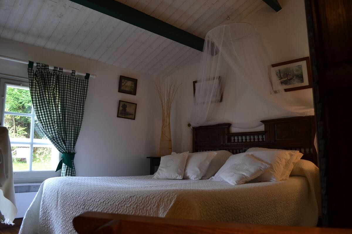 Ein Schlafzimmer mit einem Bett, einem Fenster mit Vorhängen, einem Nachttisch und einem an der Wand hängenden Bild.