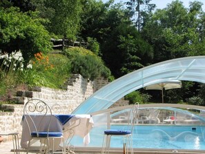 Un área de piscina con una sección envuelta en cristal, dos sillones de descanso y un muro de piedra al fondo.