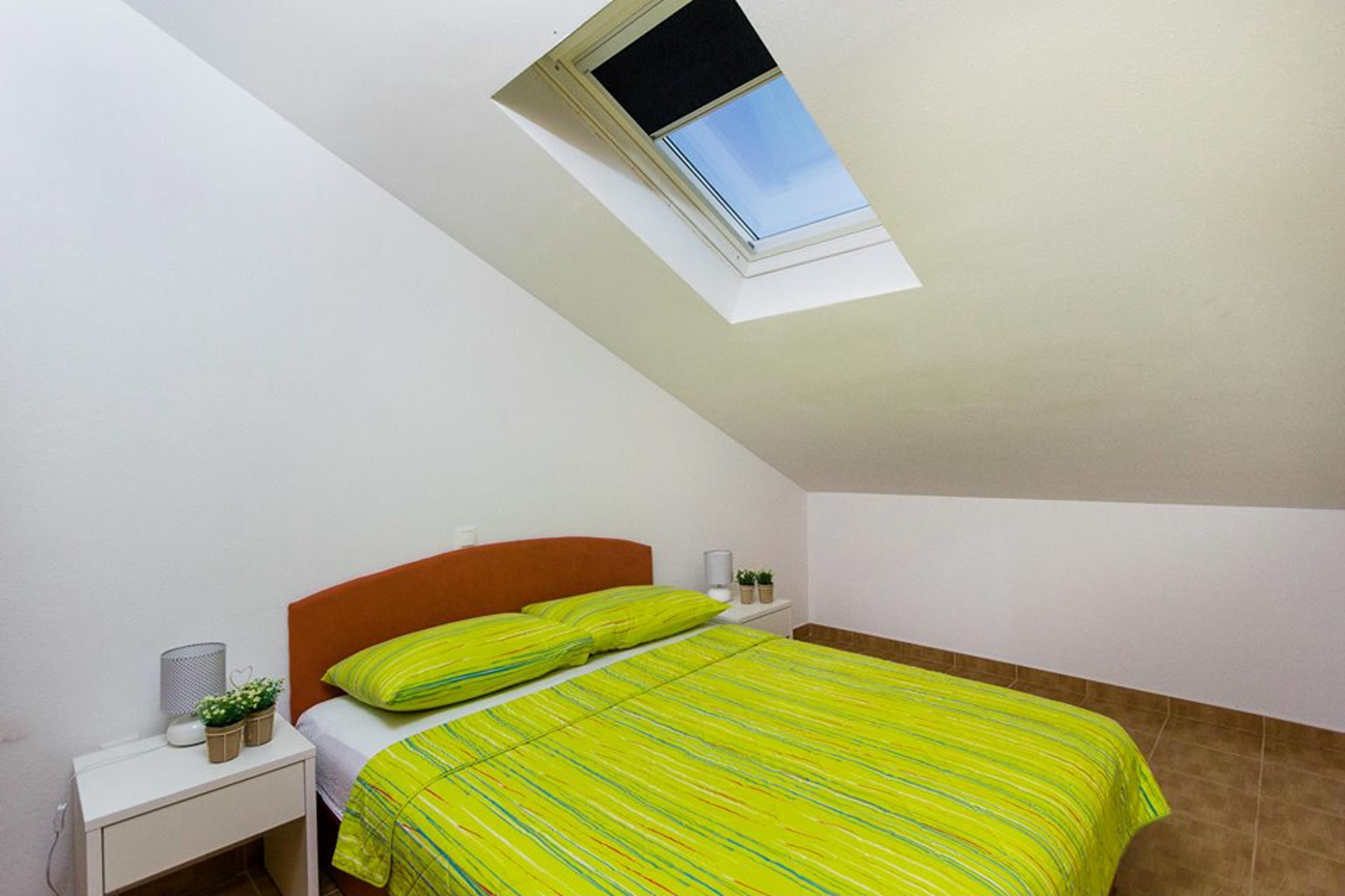 Een slaapkamer met een schuine kap, dakraam, bed met gestreept dekbed, nachtkastje met lamp en een kleine plant.