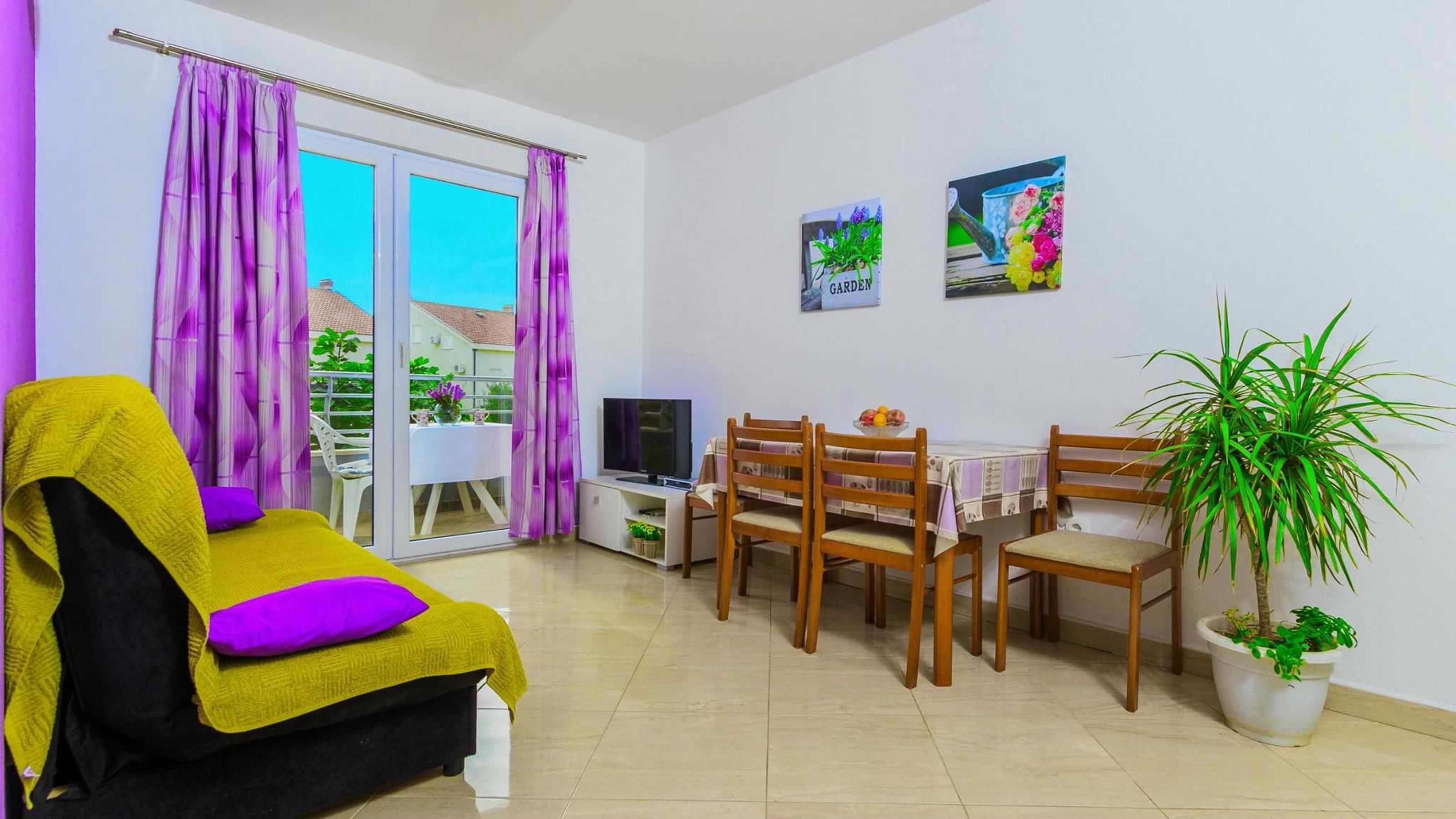 A living room with a sofa, a dining table with chairs, a TV, and a balcony with purple curtains.