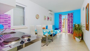 A compact kitchen with a gas stove, a sink, and a window with blinds. There is a dining area with a table and chairs, and a balcony with purple curtains. - Apartments Andrijanic (Baska Voda)
