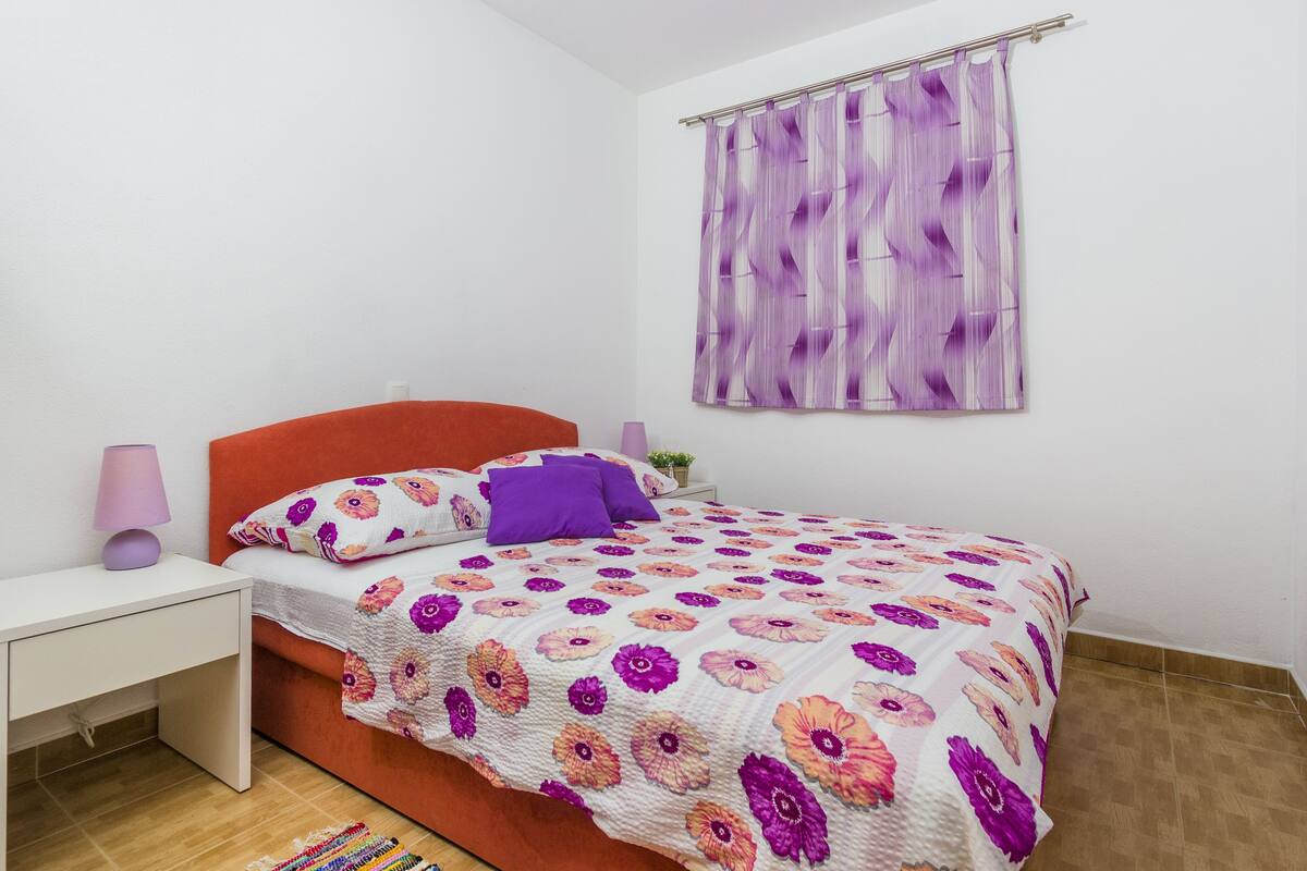 Quarto com cama, mesa de cabeceira, luminária e janela com cortina.