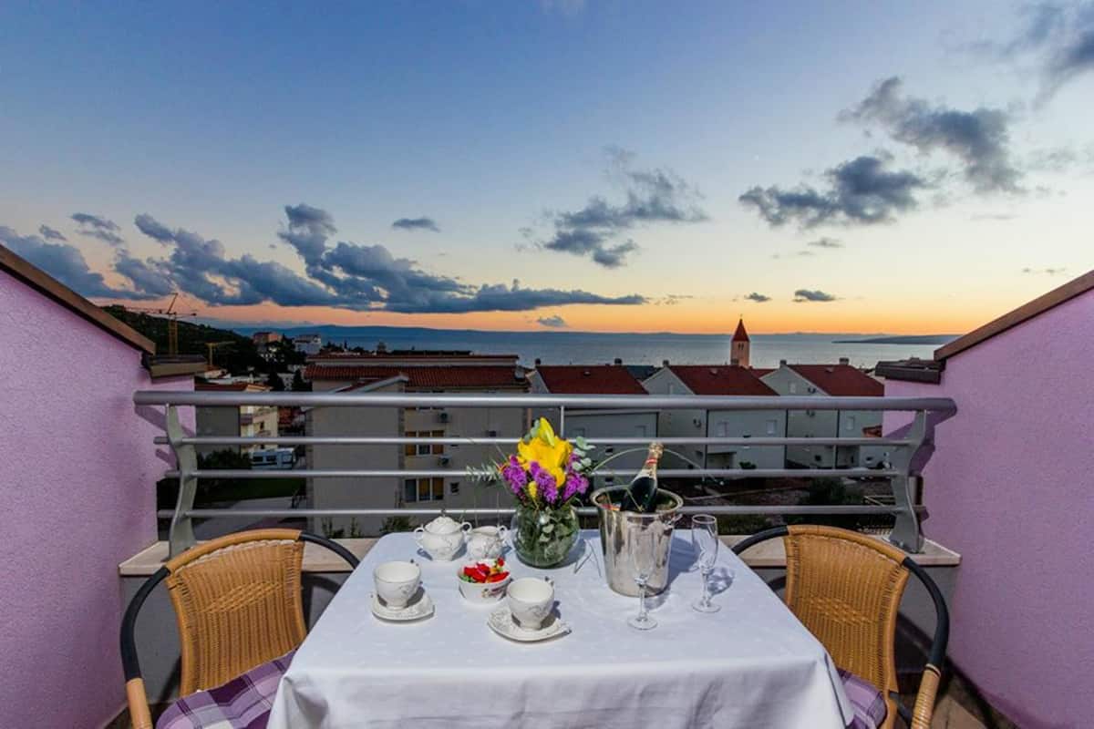 Uma varanda com uma mesa posta para o chá, um vaso com flores e vista para o mar e prédios ao pôr do sol.