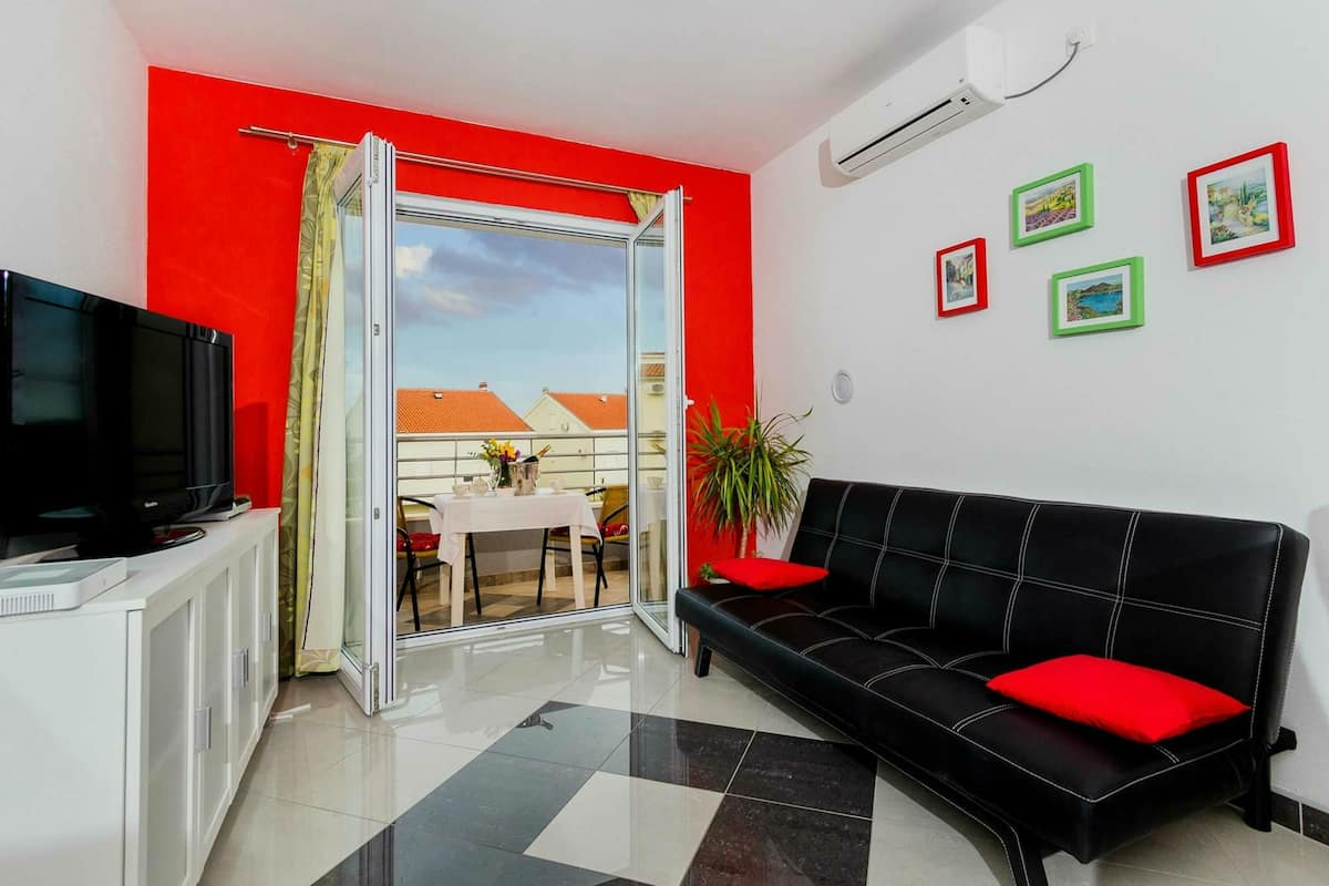 Sala de estar com sofá preto, televisão em um armário, porta de vidro deslizante para uma varanda e parede vermelha.