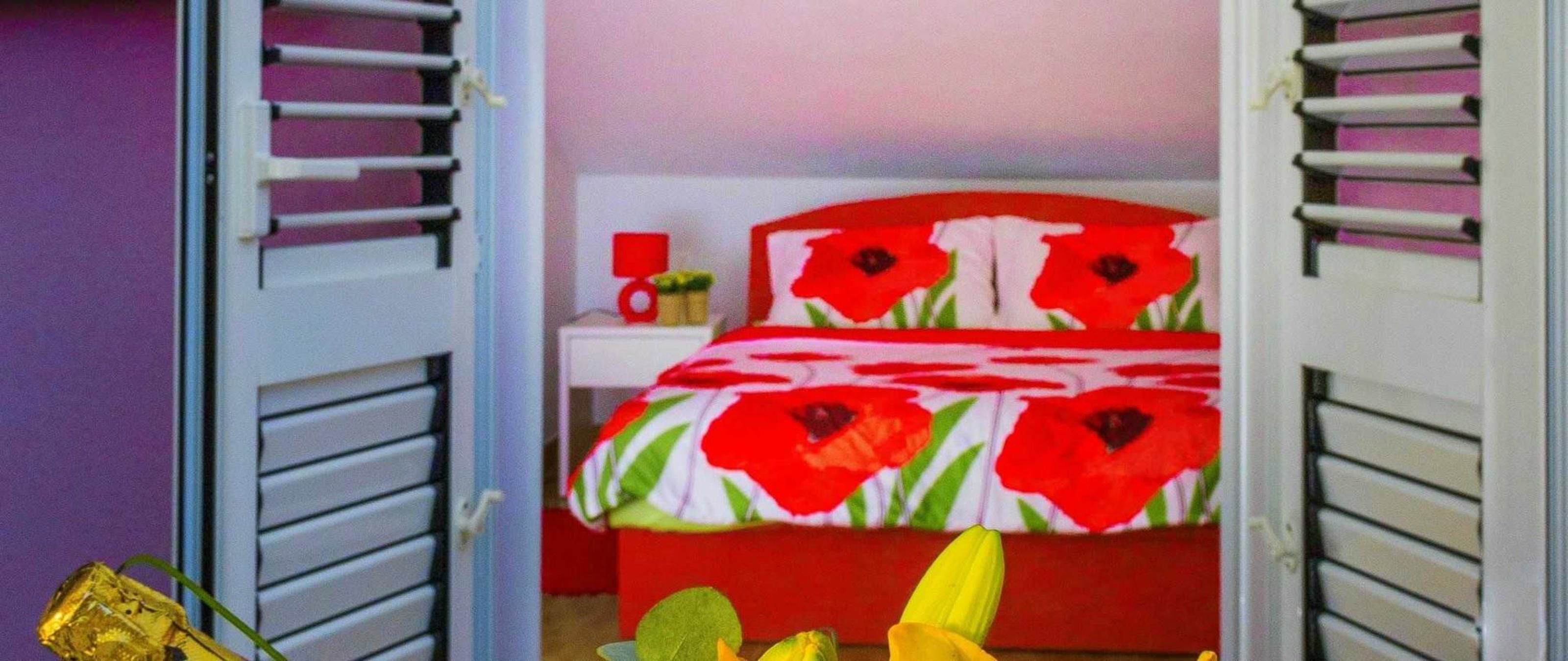 Un dormitorio con una cama con ropa de cama de flores rojas, una mesita de noche con un objeto rojo y una ventana con persianas.