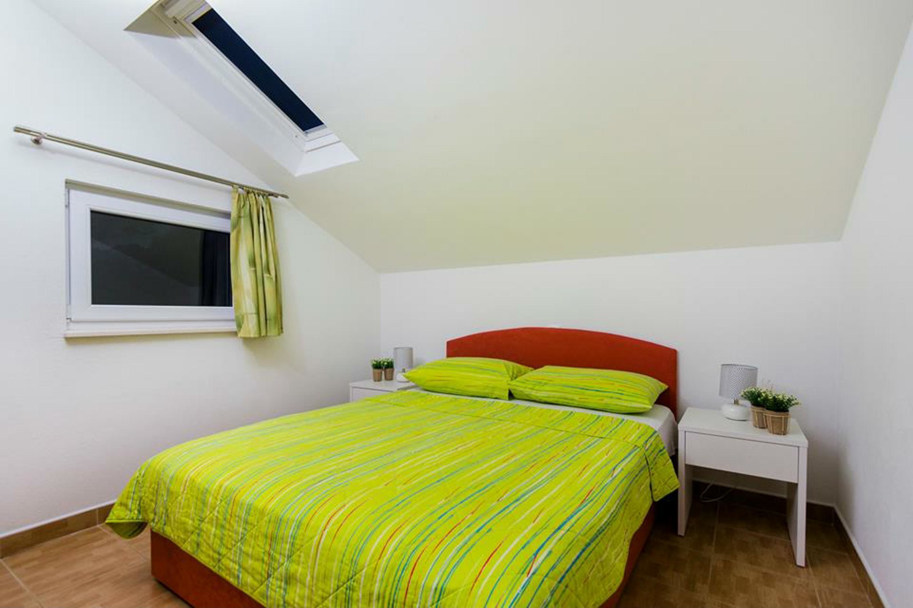 Un dormitorio con cama, una ventana con cortinas, una mesita de noche con lámpara y una planta pequeña.