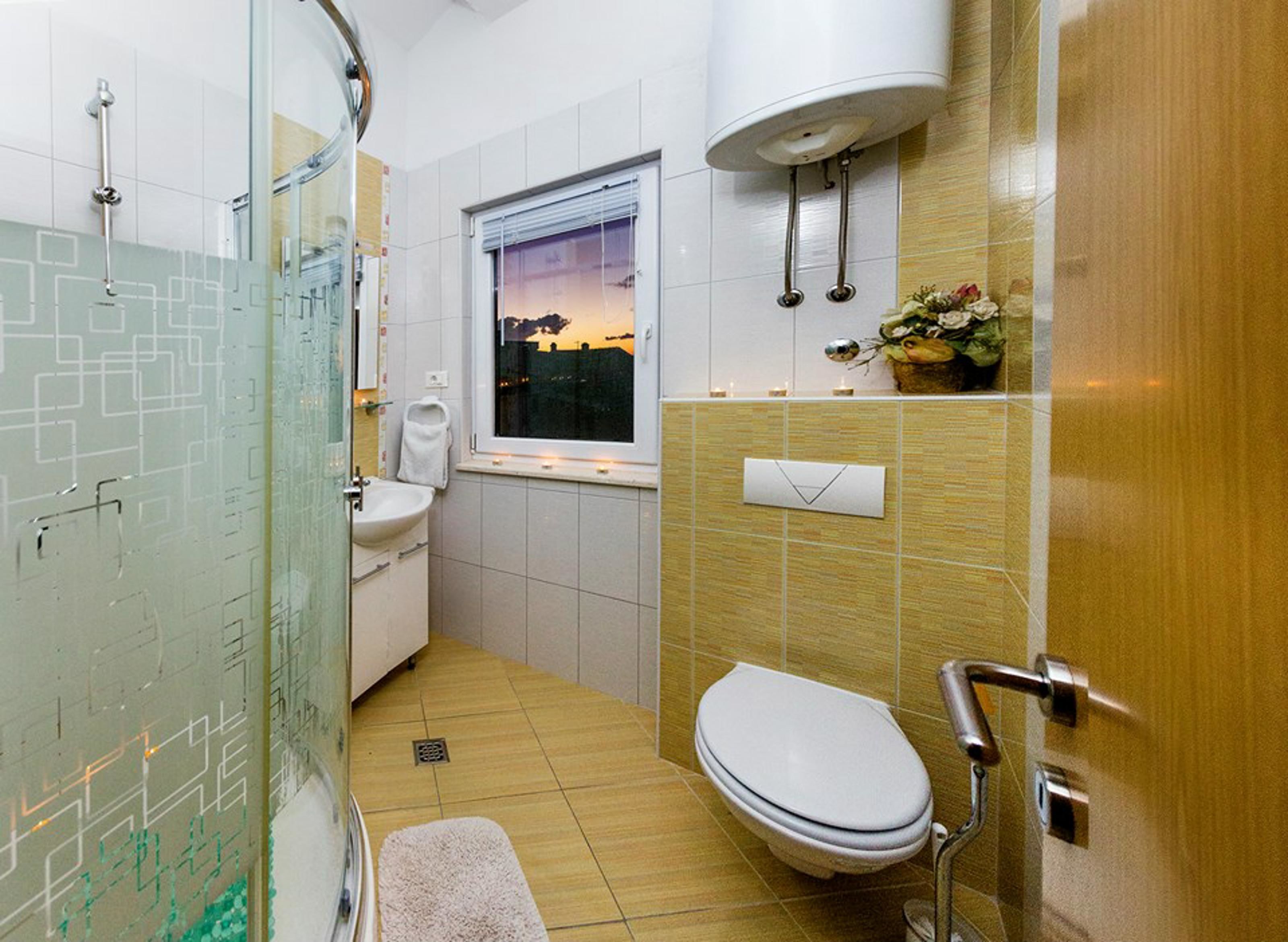 A modern bathroom with a glass shower enclosure, a toilet, a sink, and a window with a view of the sunset.