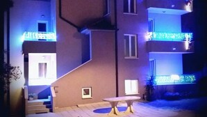 A multi-story building with balconies adorned with string lights, a staircase leading to the entrance, and a small outdoor seating area. - Apartments Andrijanic (Baska Voda)
