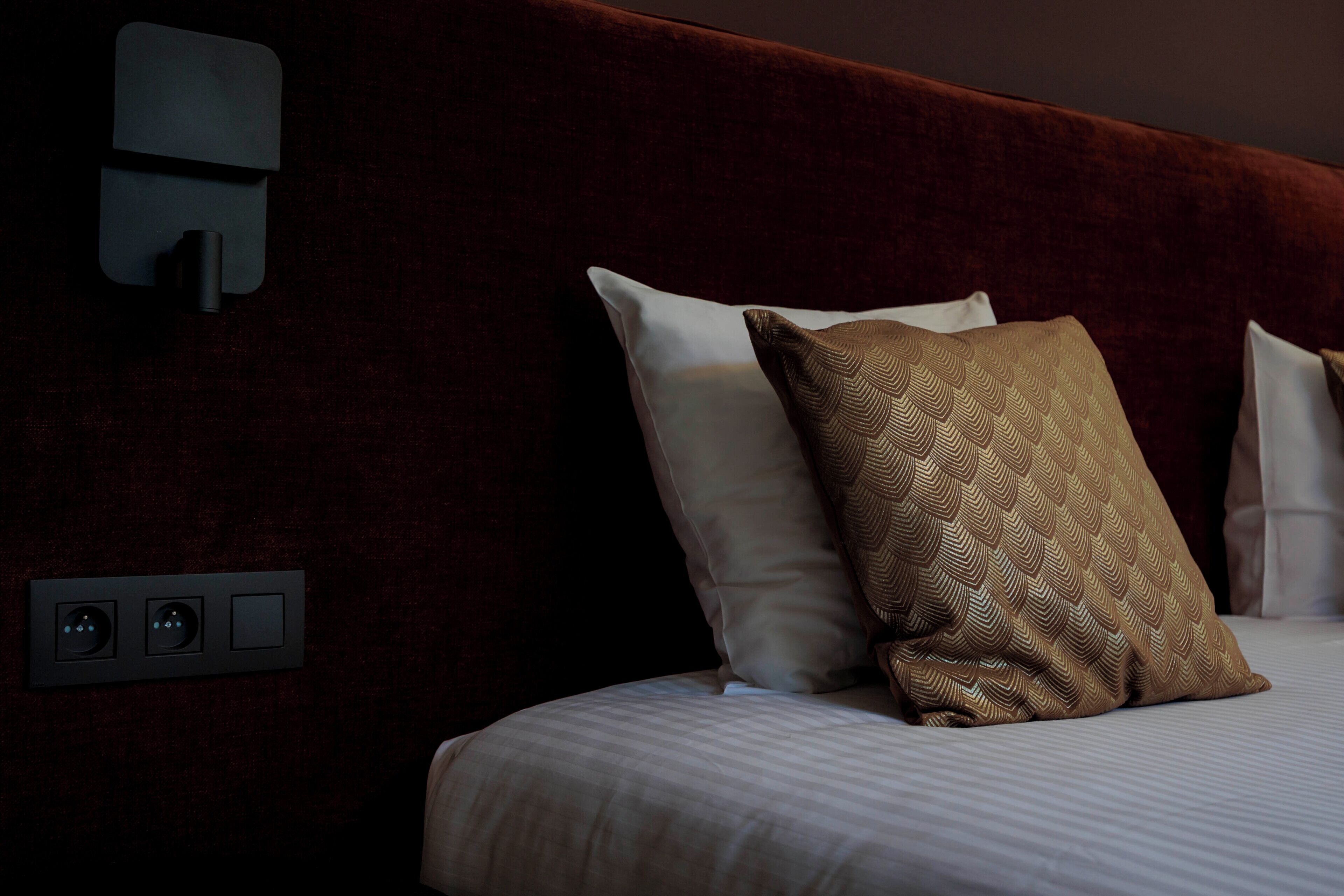 Ett hotellrum med en säng, kuddar, en väggmonterad kontrollpanel och en vägg med en intressant yta.