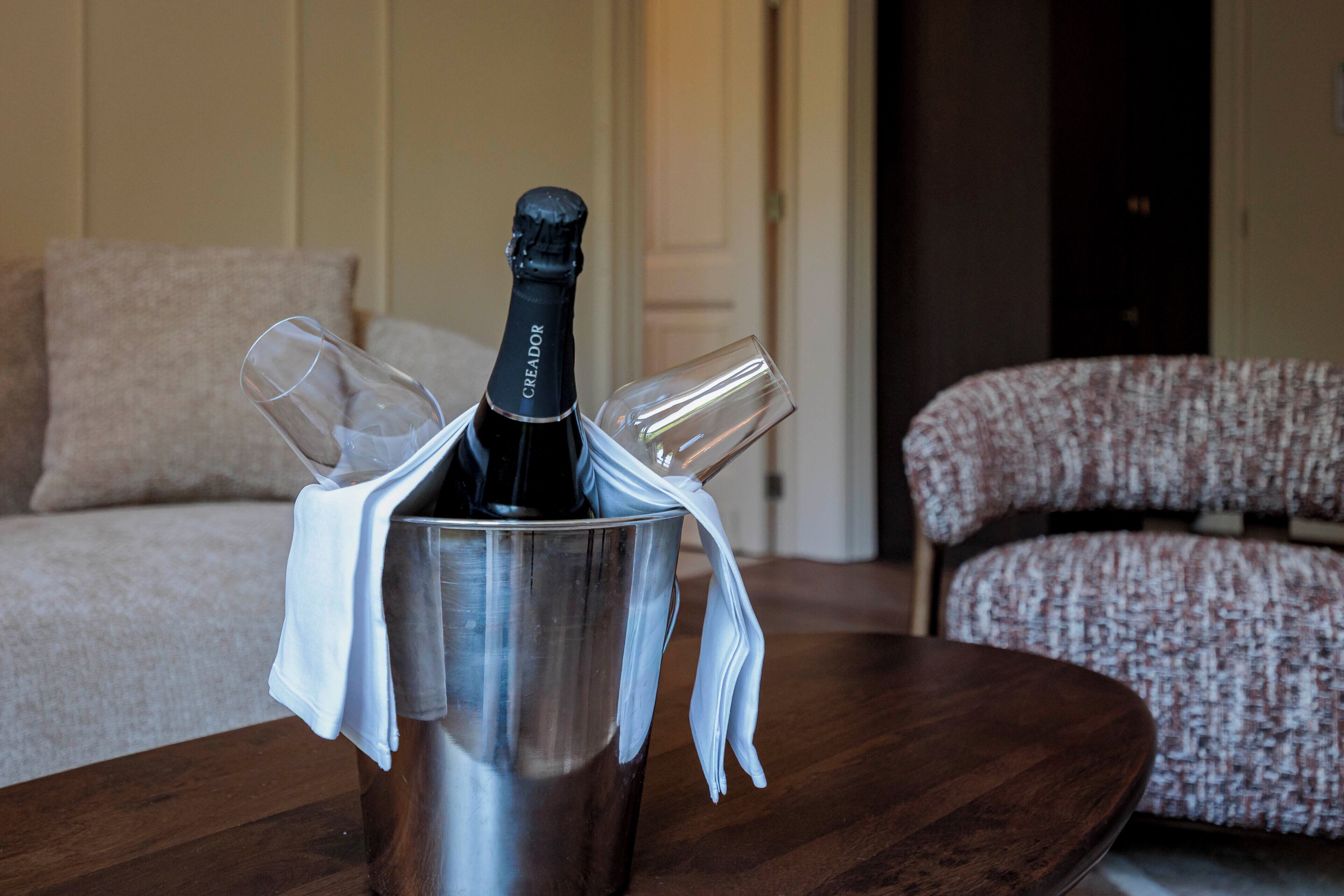 Une bouteille de vin dans un récipient en métal, accompagnée de deux verres, d’un linge blanc et d’une table en bois, le tout dans une pièce où se trouve un canapé à motifs.
