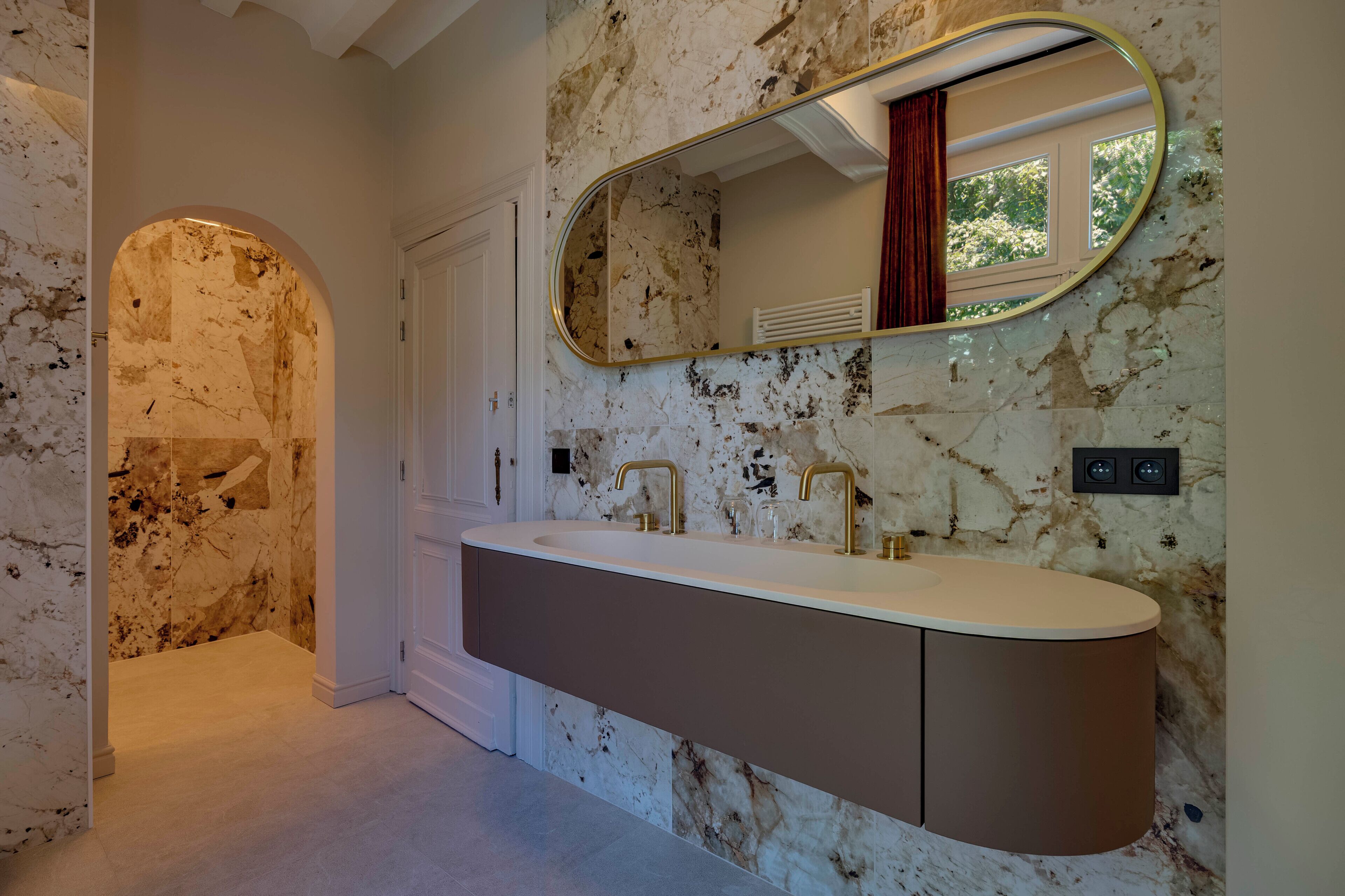 A bathroom with a marble wall, a large oval mirror, a double sink with gold faucets, and a door leading to another room.