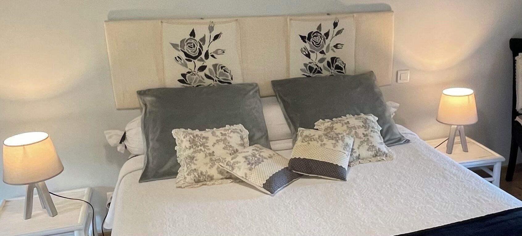 A neatly made bed with grey and white pillows, two wall-mounted lamps, and floral wall art.