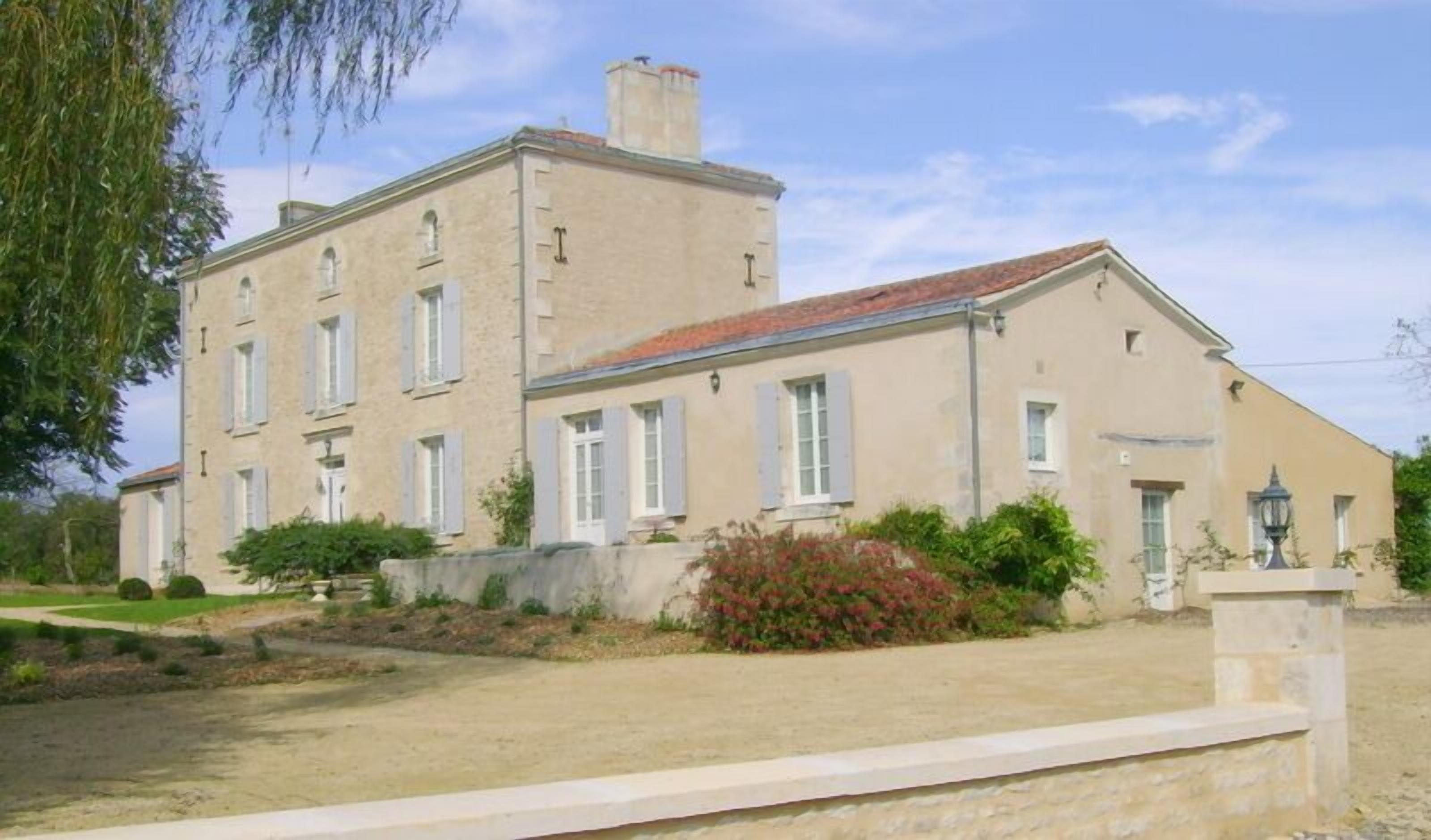 A two-story building with a red-tiled roof, multiple windows with shutters, and a well-maintained garden with shrubs and a pathway.