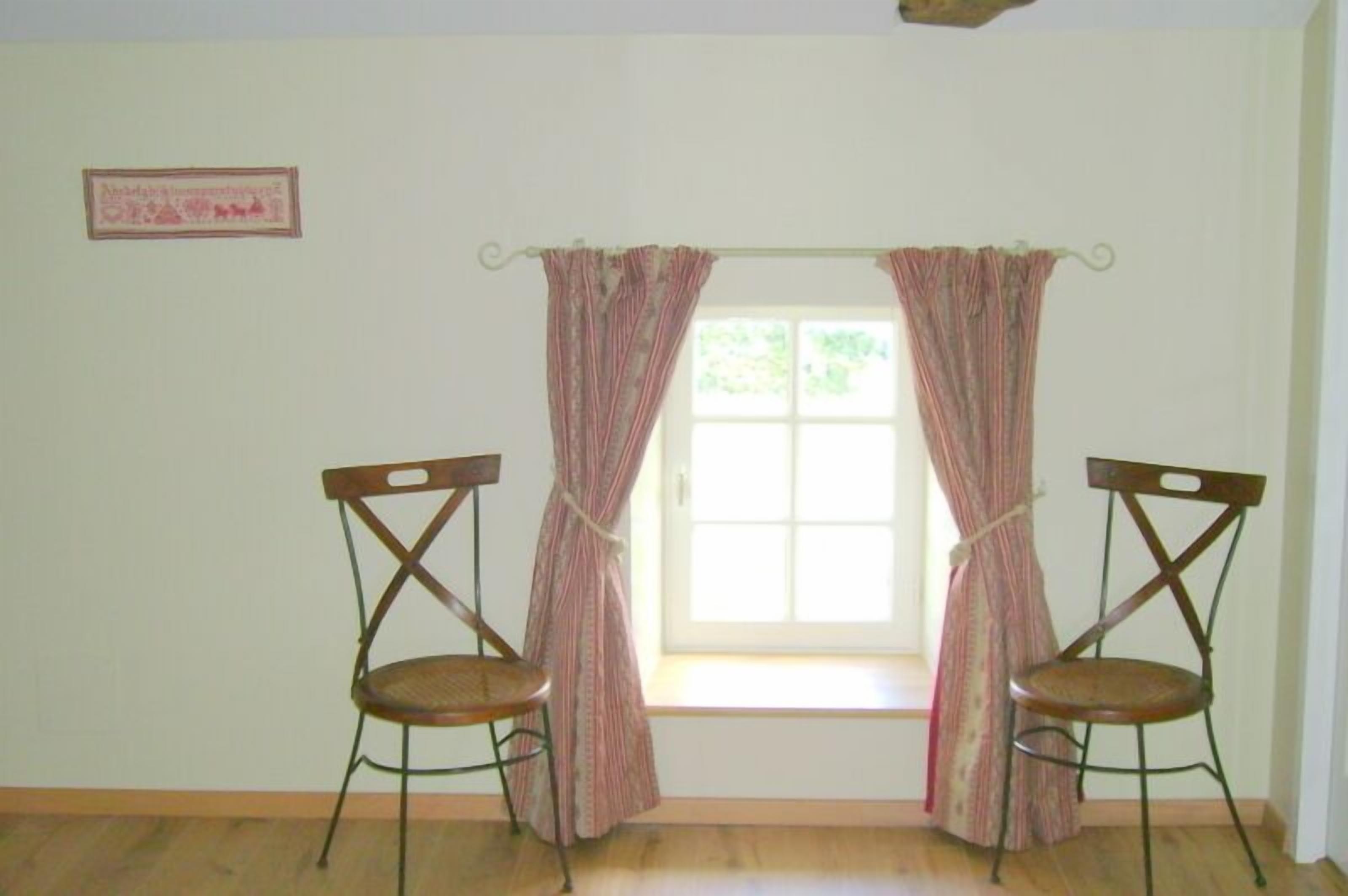 A room with two wooden chairs, a window with curtains, and a framed picture on the wall.