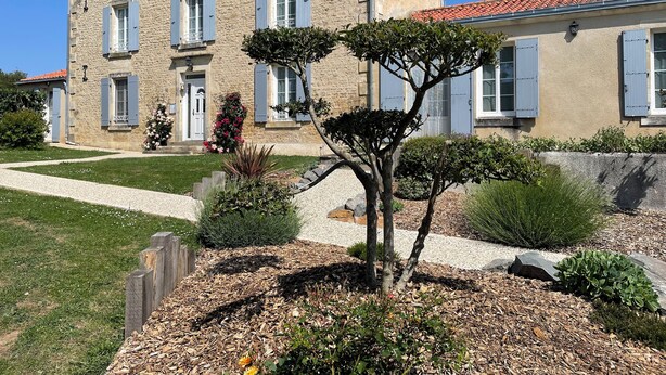 Een tweelaags stenen gebouw met blauwe luiken, een grindpad en een verzorgde tuin met verschillende planten en bloemen.