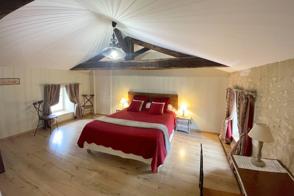 Un dormitorio con una cama grande, vigas de madera, un ventanal con cortinas, una silla y una mesita de noche con una lámpara.