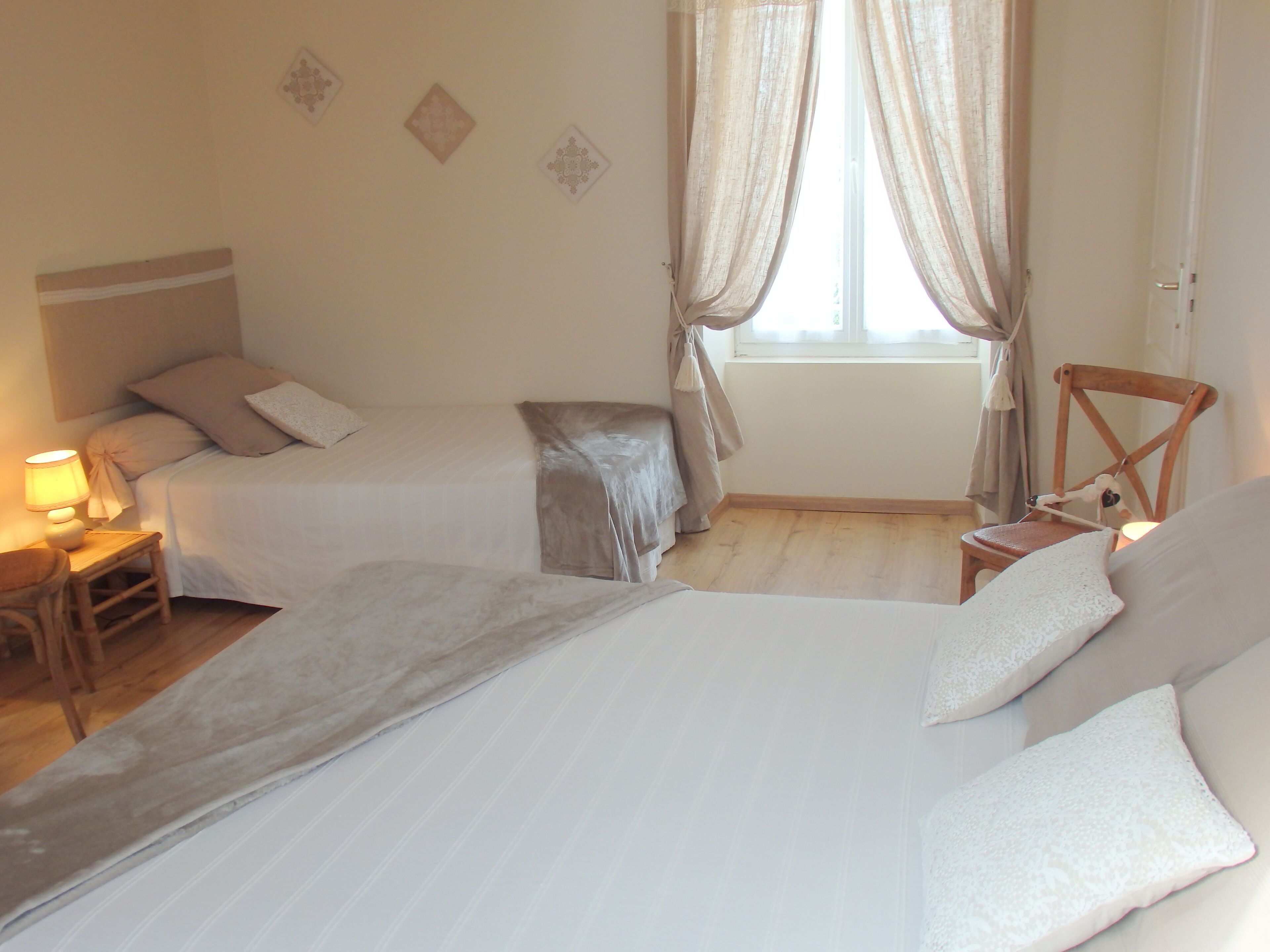 Un dormitorio con dos camas, una mesita de noche con lámpara, una silla y un ventanal con cortinas.