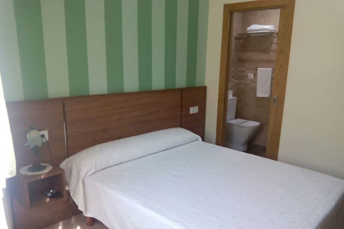 A bedroom with a wooden headboard, a single bed with white bedding, a nightstand with a lamp, and a bathroom visible through an open door.