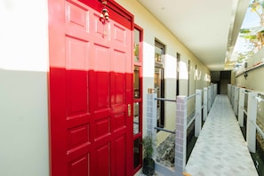 A red door with a small potted plant beside it, a tiled walkway, and a view of a building in the background. - RedDoorz Plus @ Ruby Las Pinas Weekly Stay (Las Pinas)