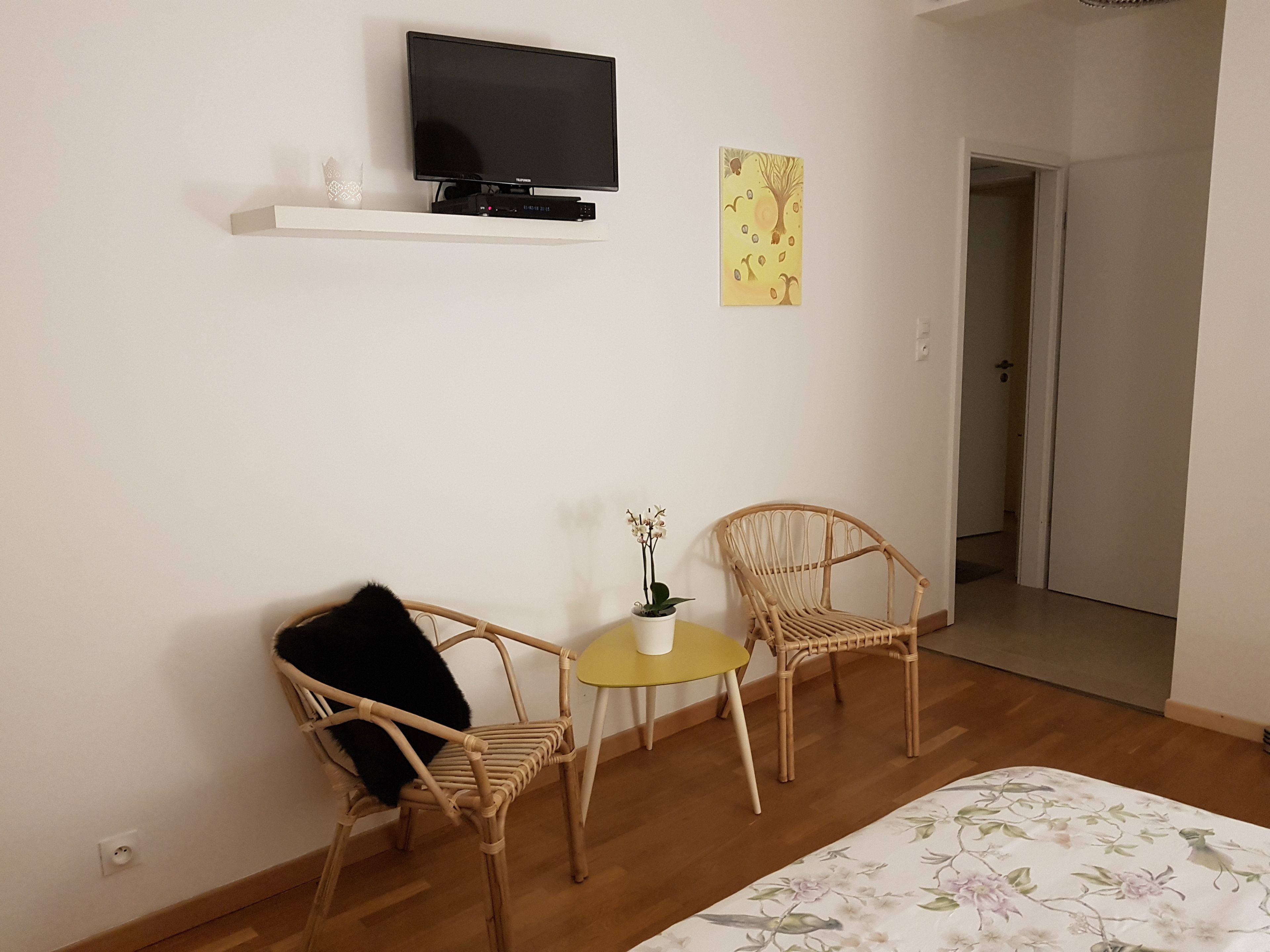 Una habitación con un televisor en un estante, dos sillas de mimbre, una mesita con una planta y un edredón estampado de flores.