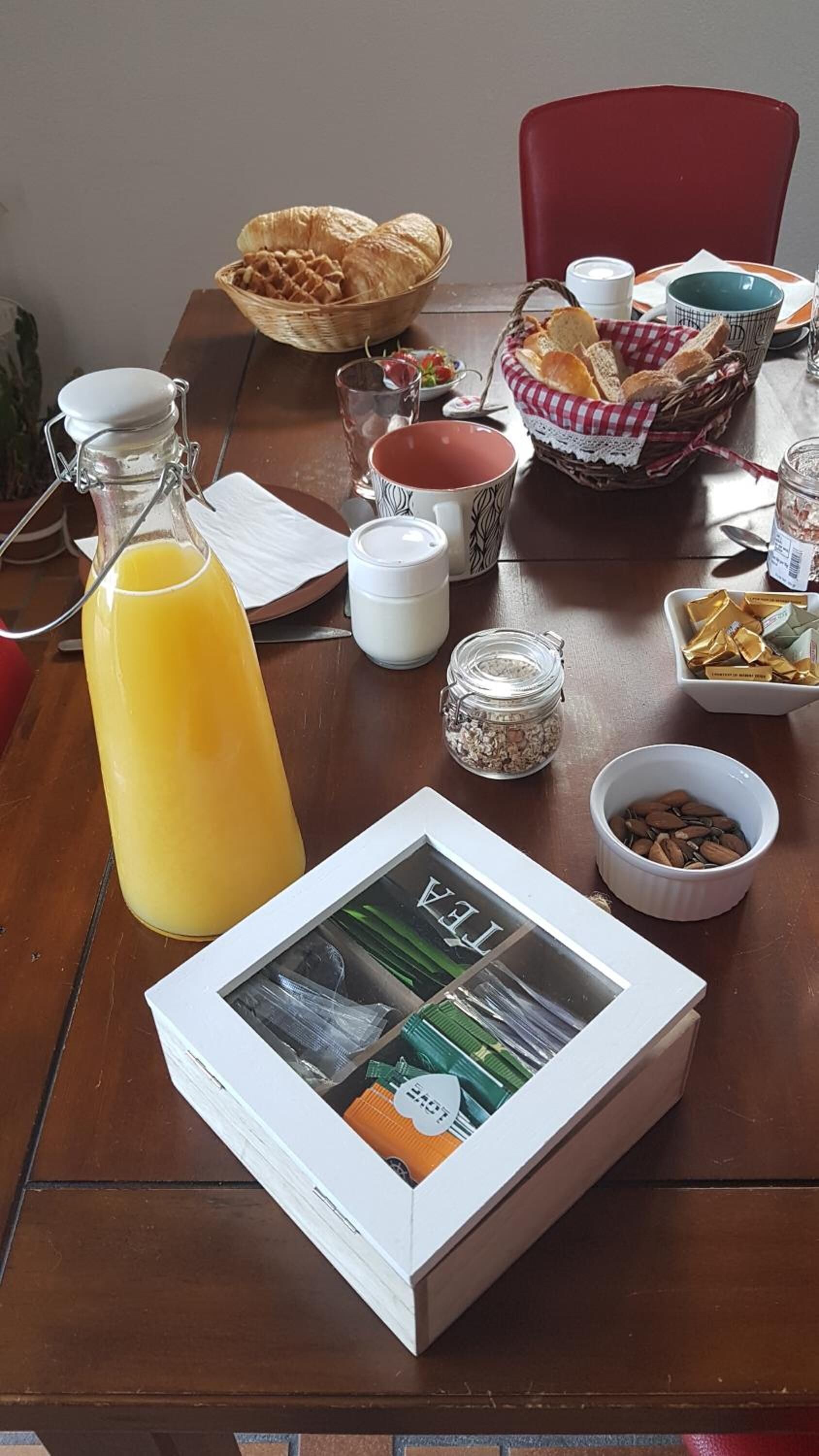 A breakfast table with a bottle of orange juice, a glass, a basket of bread, a bowl of almonds, and a box of tea.